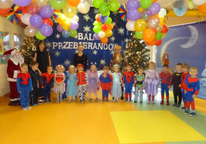 Grupa dzieci stoi z panią dyrektor Marią Królikowską, p. Anią i p. Natalią na tle dekoracji, pomiędzy przystrojonymi choinkami.